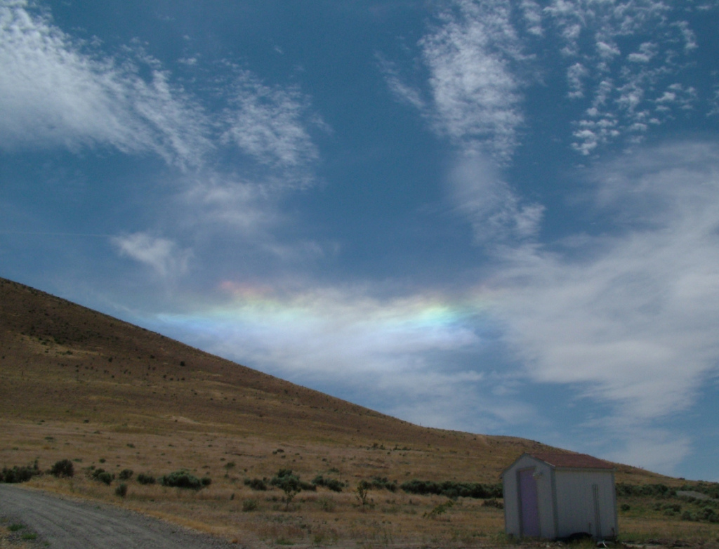 A patch of iridescent clouds in rainbow colors, in a blue partly-cloudy sky above a brown arid hillside.